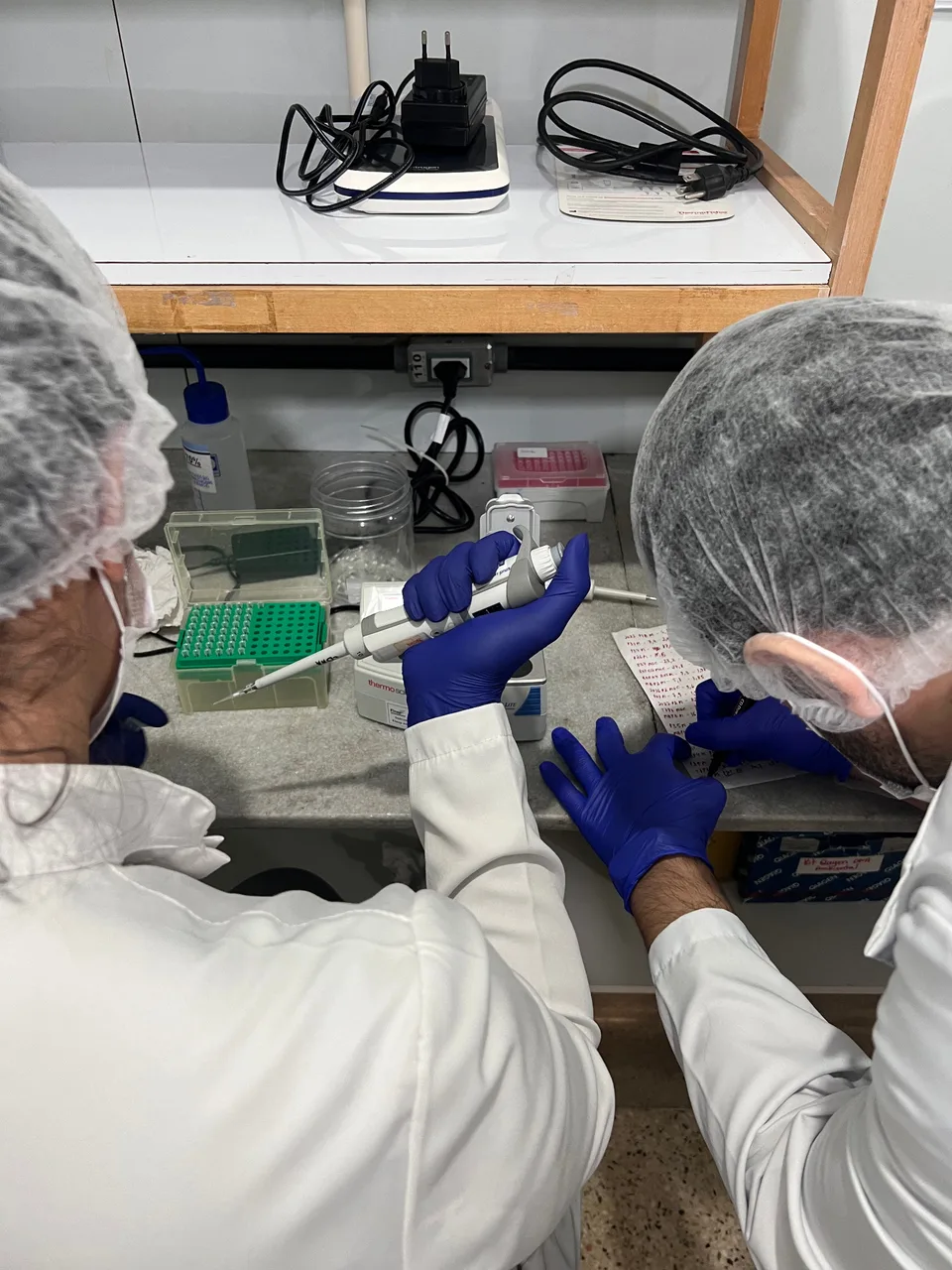 Duas pessoas com equipamentos de proteção trabalhando em uma bancada de laboratório.